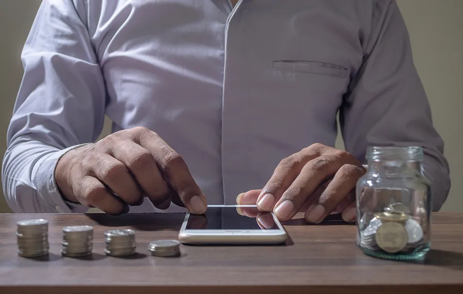 man touching a mobile phone on a desk with coins in a jar and stacked on a table