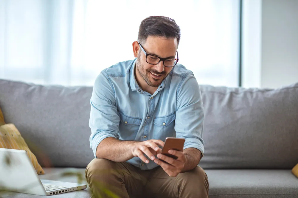man with glasses and blue button up shirt sitting on a grey couch smiling at a mobile phone in his hands