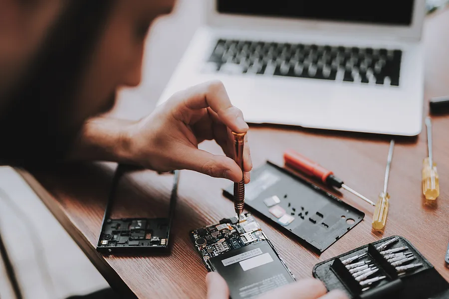 A technician uses a precision screwdriver to repair the internal components of a smartphone on a wooden desk, surrounded by small tools, spare parts, and an open laptop.